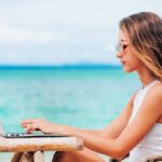 Young woman freelancer in dress sitting at the table on ocean background, using laptop on the beach. Girl Freelancer working