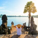 One tourist relaxing in Angkor ruins at sunrise, Srah Srang temple water pond amid jungle, travel destination Cambodia. Woman with traditional hat, rear view.