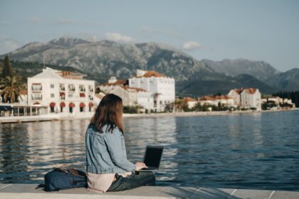 Young Woman Digital Nomad Working From Her Laptop As She Sits On A Boardwalk In A Coastal Town In Montenegro, Balkan Peninsula, South Eastern Europe